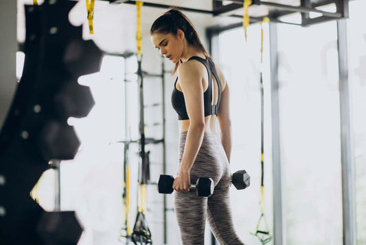 Woman using dumbbells in gym for functional fitness workout with resistance bands and kettlebells in background.