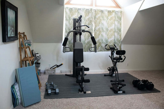 Home gym setup with affordable equipment including a multi-gym machine, dumbbells, and exercise bike in a carpeted room.