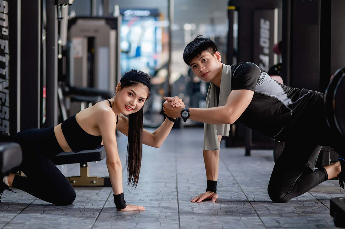 Two people exercising together in a gym setting with various gym equipment in the background.