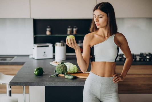 Woman in workout gear holding an apple, surrounded by vegetables, symbolizing healthy eating and fitness choices in a modern kitchen.