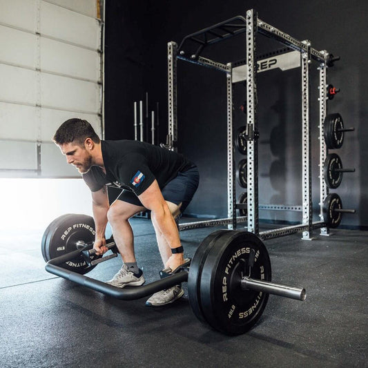 Man using open trap bar for strength training in a home gym, featuring gym equipment like weights and racks.